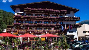 Ein mehrstöckiges Hotel mit Balkonen, die mit Blumen geschmückt sind, Sitzgelegenheiten im Freien mit roten Sonnenschirmen und Flaggen, die internationale Auszeichnungen kennzeichnen.