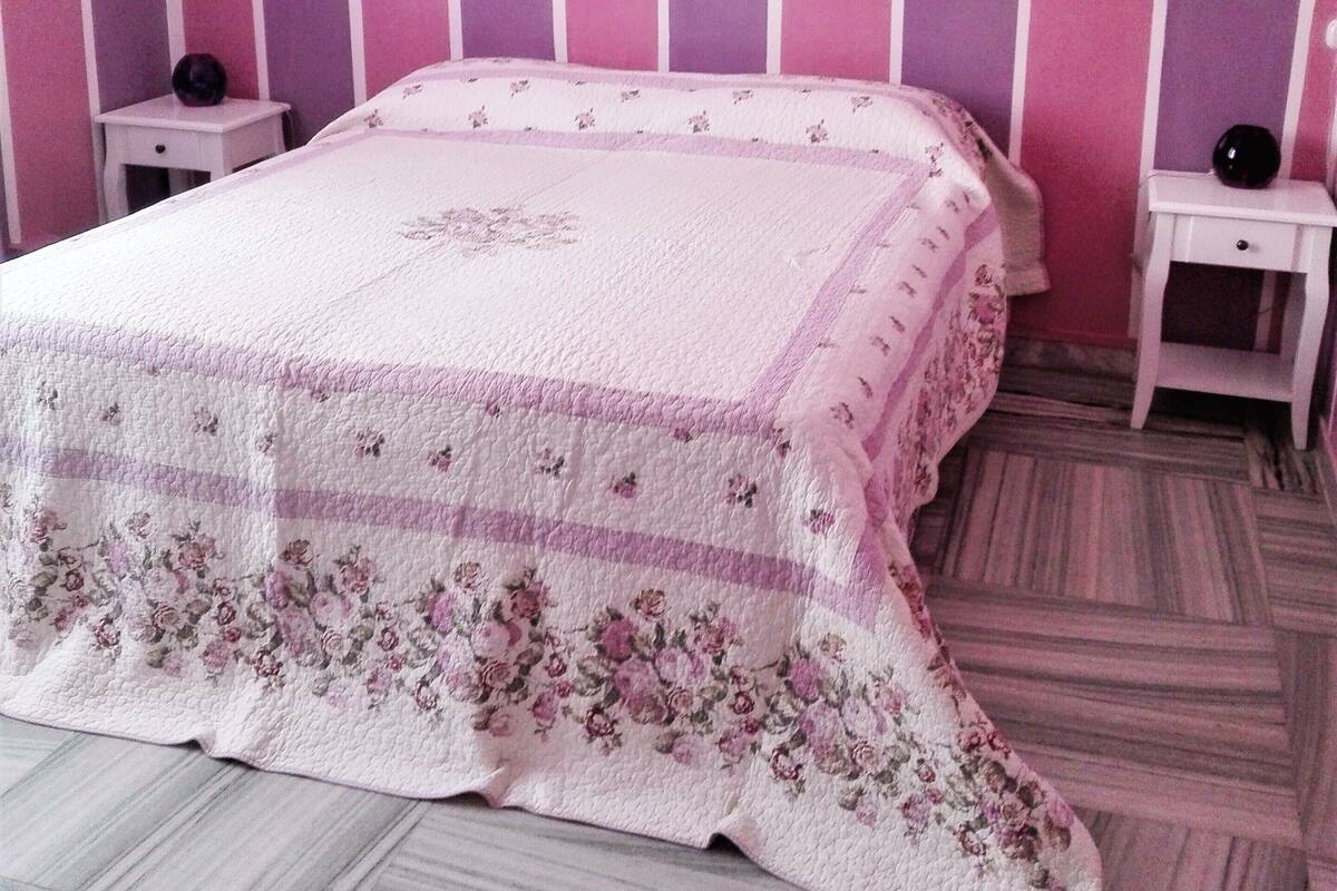 A bedroom with a pink and white color scheme, featuring a bed with floral bedding, two bedside tables, and a wall-mounted artwork of flowers.