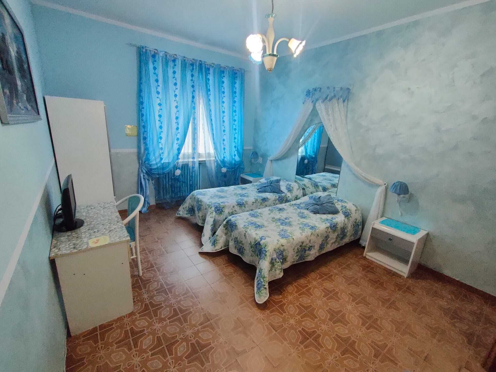 A bedroom with two beds, a desk, a chair, a bedside table, and a window with curtains.