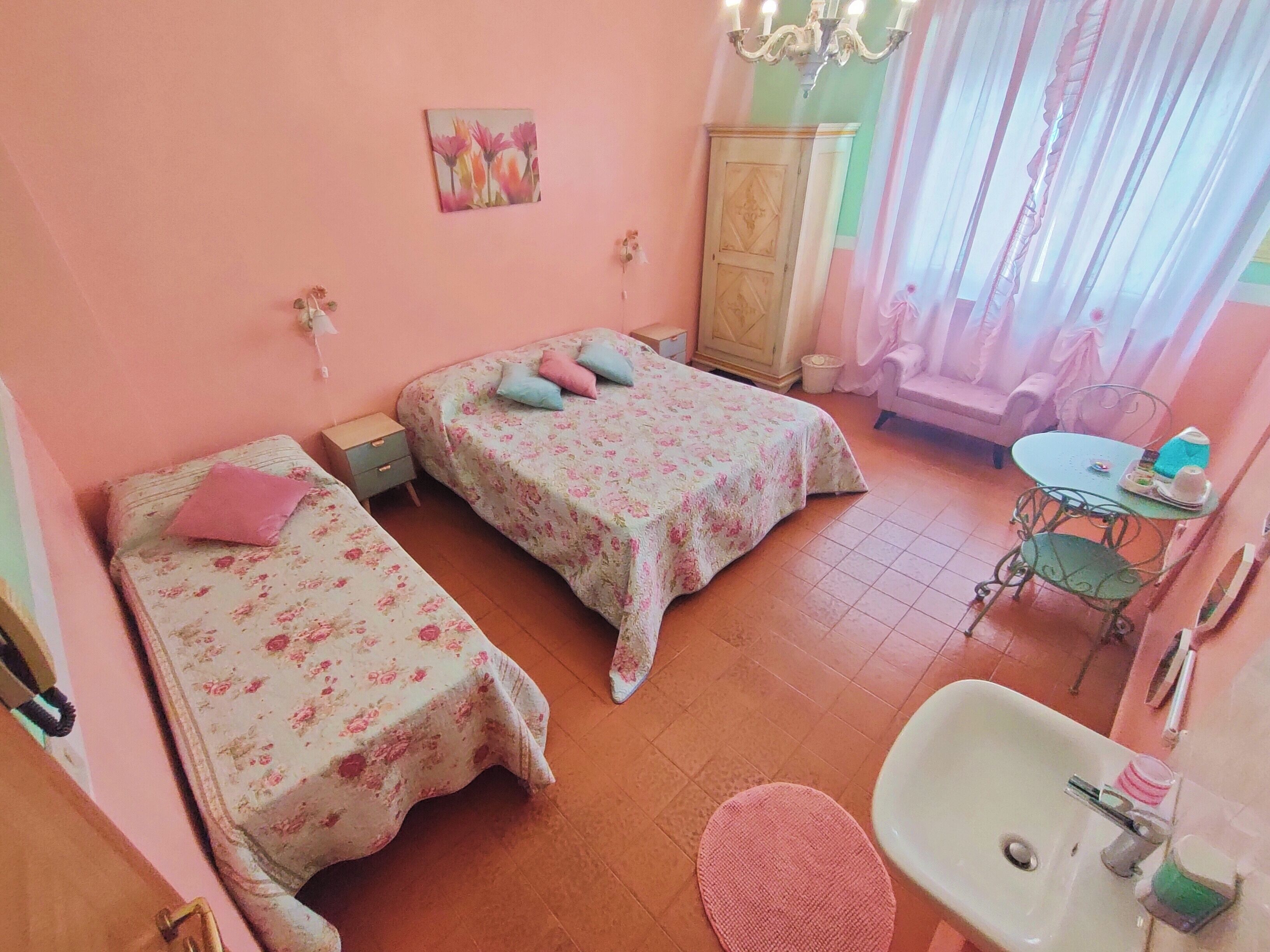 A bedroom with two beds, a wardrobe, a chandelier, a table with a chair, and a sink.