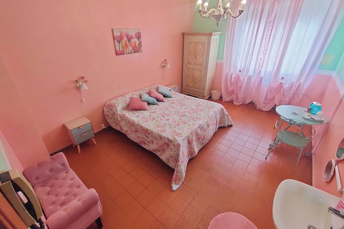 A bedroom with a floral bedspread, a pink armchair, a small desk with a chair, a wardrobe, and a chandelier.