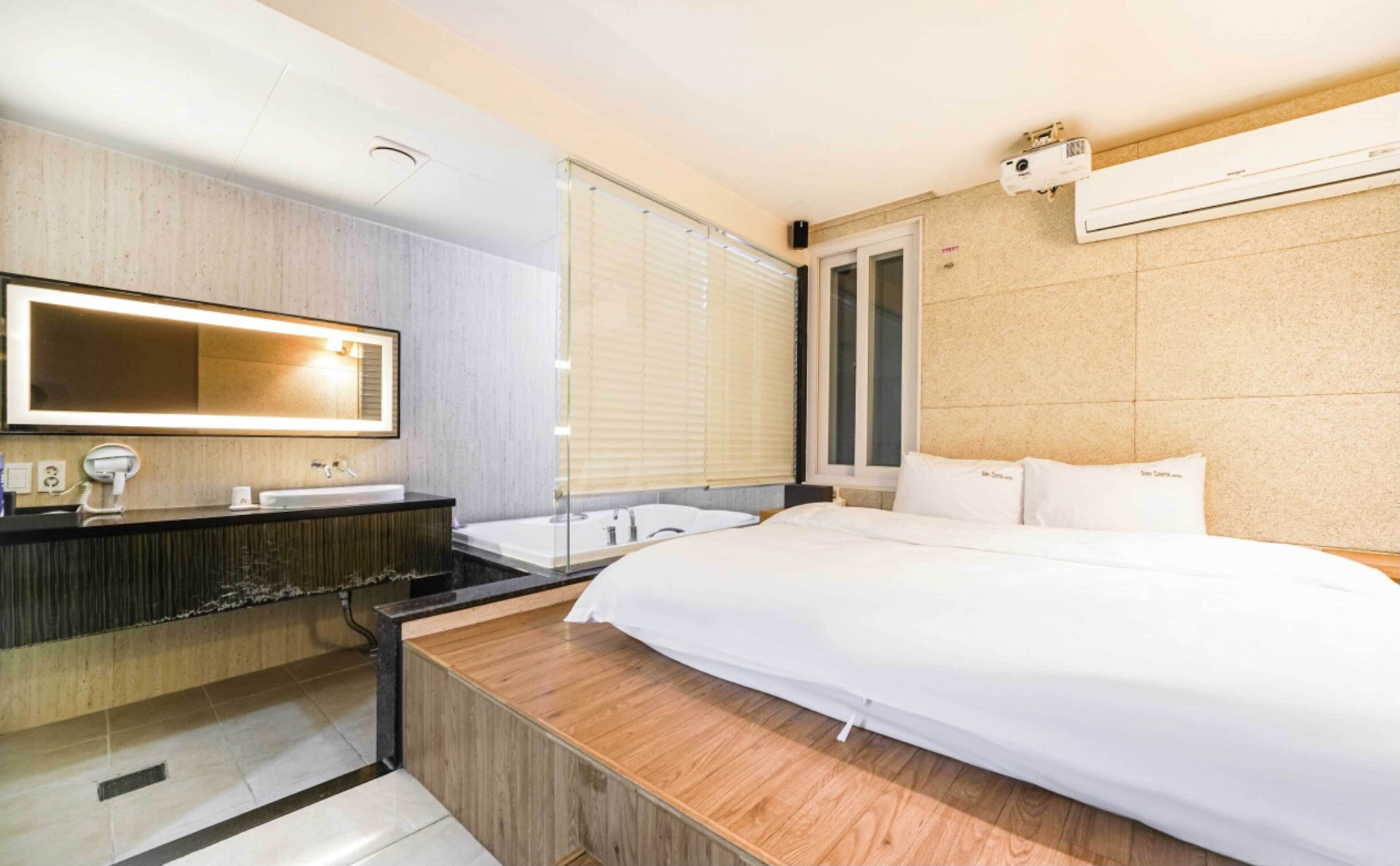 Habitació d'hotel moderna amb un llit gran, una taula de nit de fusta, un tocador integrat amb mirall, una banyera i un aparell d'aire condicionat a la paret.