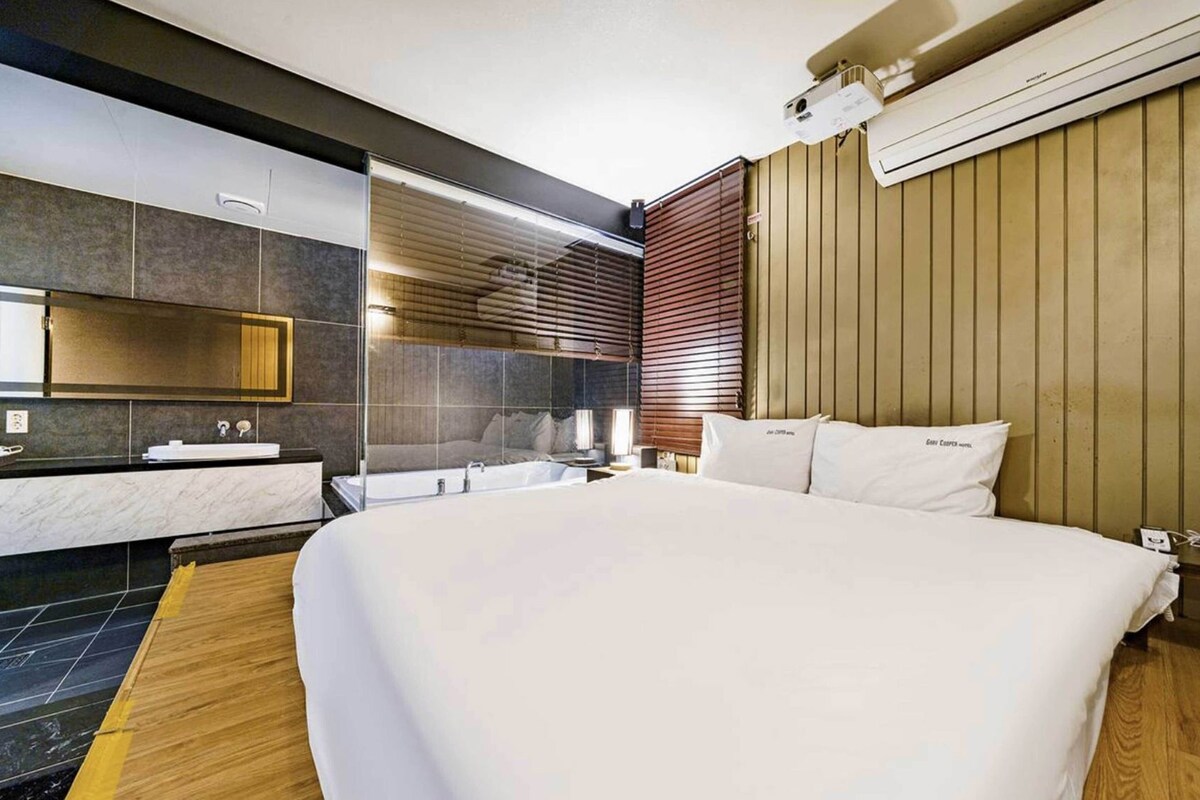 A modern hotel room with a large bed, a bathroom with a mirror and sink, and a wooden panel wall.