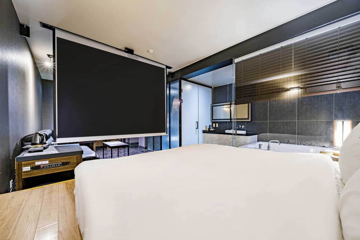 Moderni hotellihuone, jossa on suuri sänky, työpöytä kannettavalla tietokoneella, taulutelevisio, kylpyhuone, jossa on suuri peili, ja näkymä kaupunkiin lattiasta kattoon ulottuvista ikkunoista.