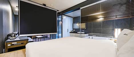 A modern hotel room with a large bed, a desk with a laptop, a flat-screen TV, a bathroom with a large mirror, and a view of the city through floor-to-ceiling windows.