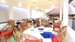 A dining area with round tables and wicker chairs, featuring a view of a building with a red roof. - New Agena Hotel (Bujumbura)