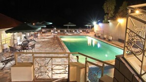 A nighttime view of a pool area with lounge chairs and umbrellas, surrounded by a stone-tiled deck and a metal railing. - New Agena Hotel (Bujumbura)
