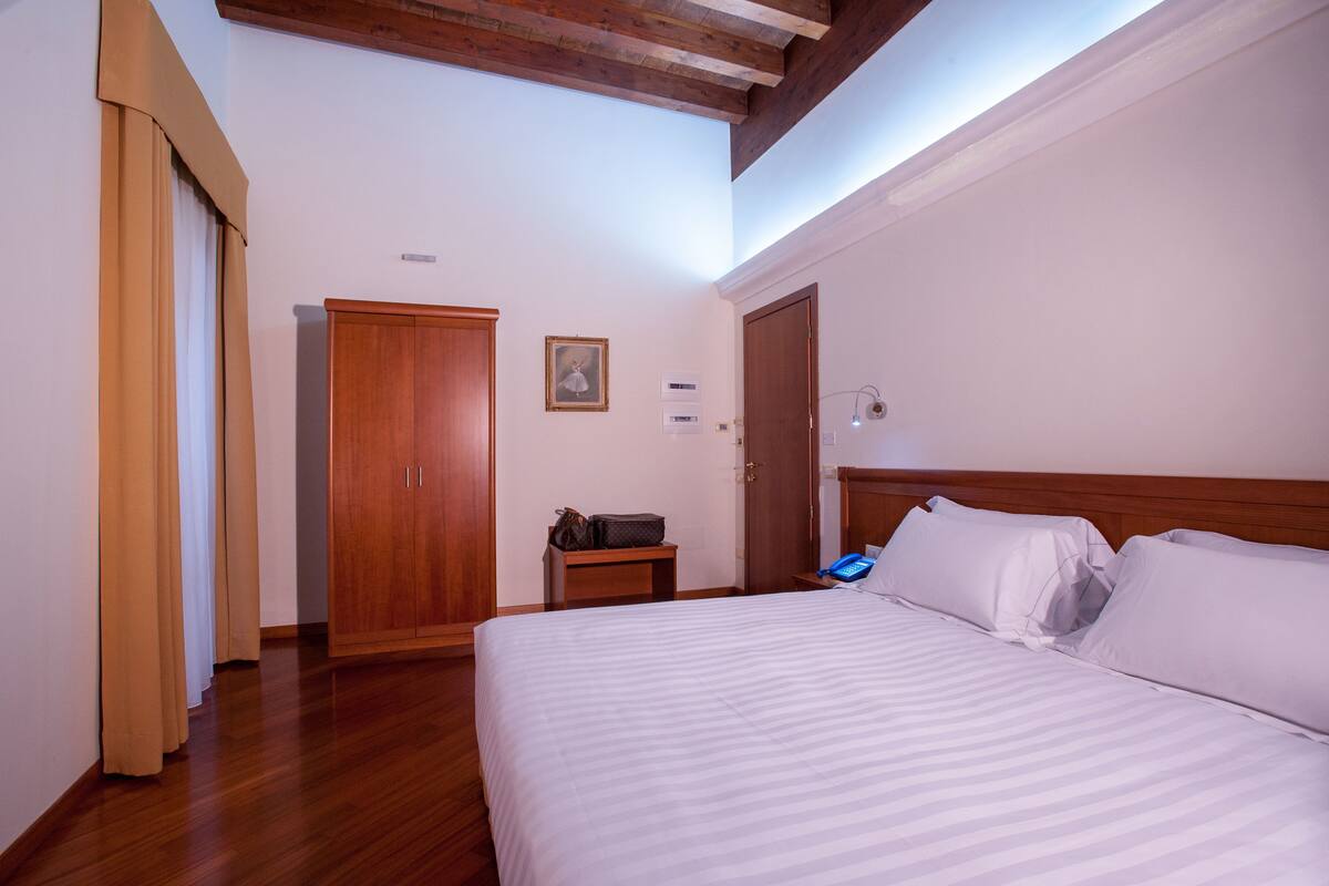 A hotel room with a large bed, wooden wardrobe, and a small bedside table with a lamp.