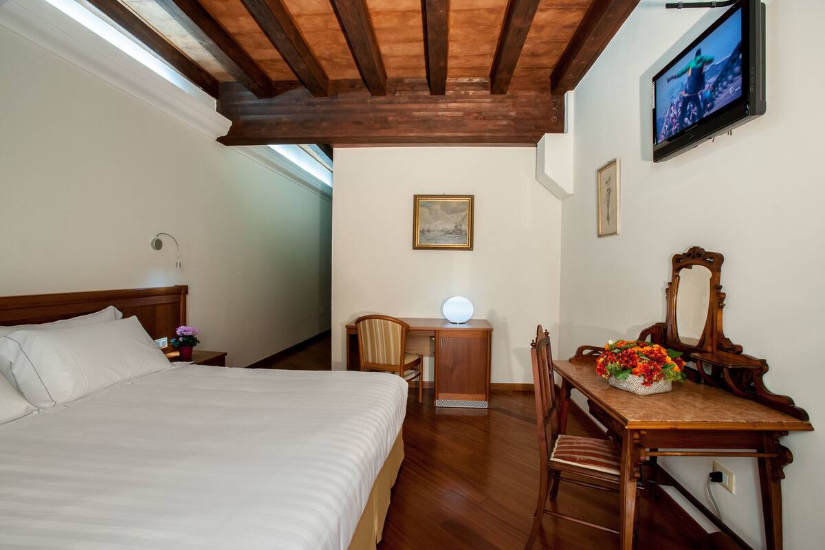 A hotel room with a bed, a desk with a chair, a desk lamp, a mirror, and a television on the wall.