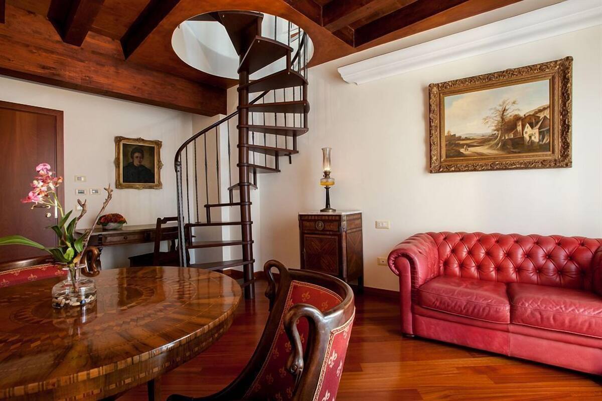 A dining area with a wooden table, chairs, a red leather sofa, a spiral staircase, and a framed painting on the wall.