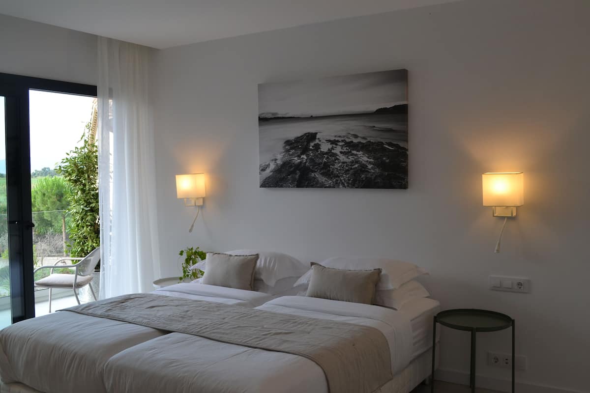 Ein Schlafzimmer mit einem Bett, zwei Nachttischlampen, einem Bild und Blick auf Grün durch ein Fenster.