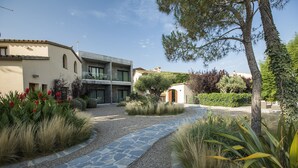 Un patio con un camino de piedra, rodeado de diversas plantas y árboles, que conduce a un edificio con múltiples ventanas y balcones.
