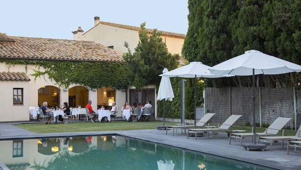 Un área de piscina con sillones de relax y sombrillas, una zona de comedor con mesas y sillas, y un edificio con tejado de baldosas y vegetación.