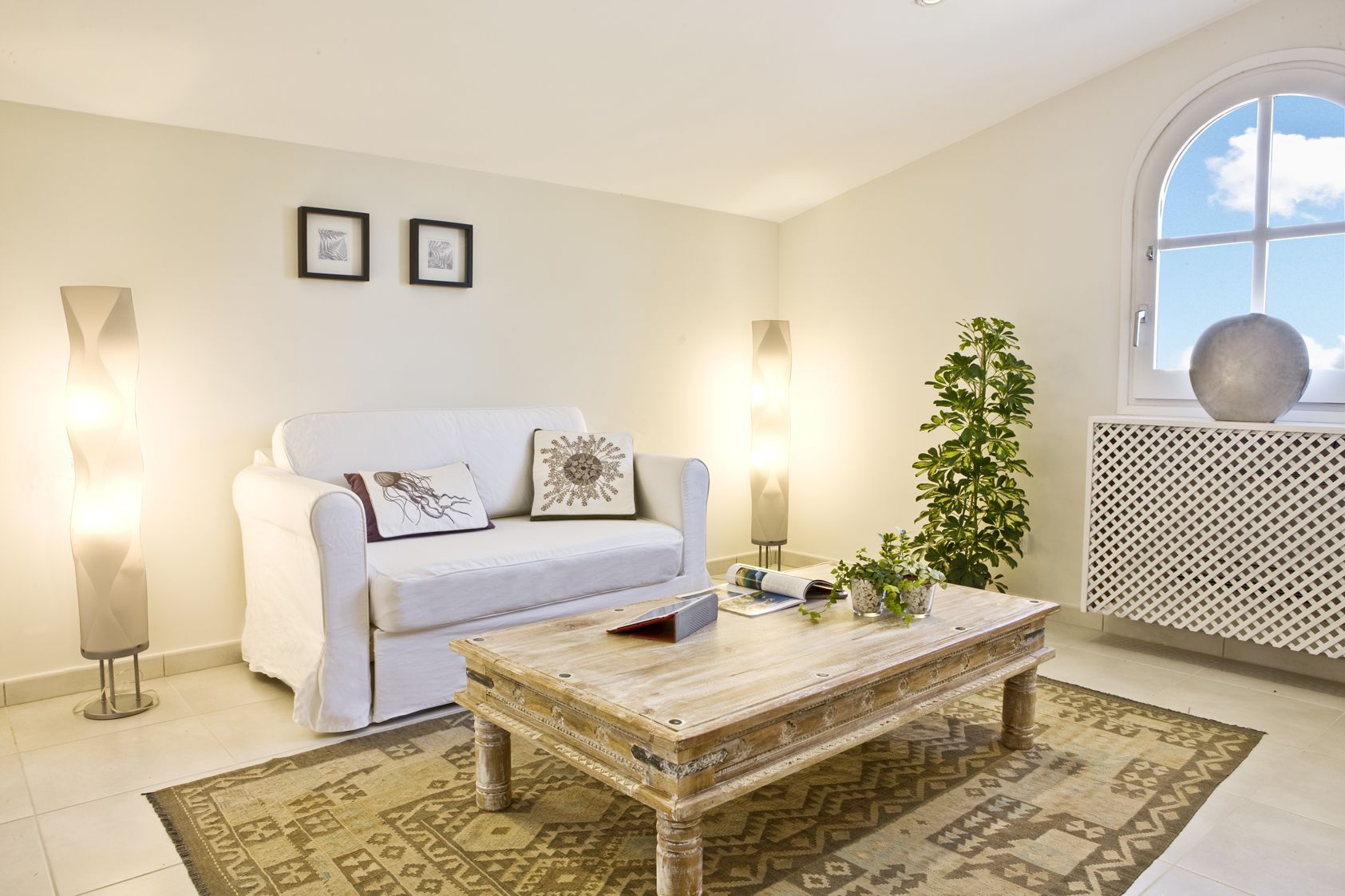 Un salón con un sofá blanco, dos cuadros enmarcados, una mesa de centro de madera, una alfombra estampada, una planta en maceta y un amplio ventanal con vistas.