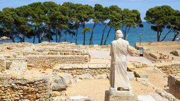 Une ruine antique, avec des structures en pierre et une statue, entourée d’arbres et de la mer.