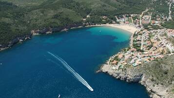 An aerial view of a coastal town with a clear blue bay, a winding road, and a boat creating a wake in the water.