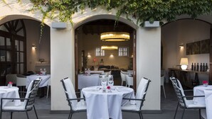 Un espacio de comedor al aire libre con mesas y sillas redondas, cubiertas con manteles blancos, y una lámpara colgante arriba.
