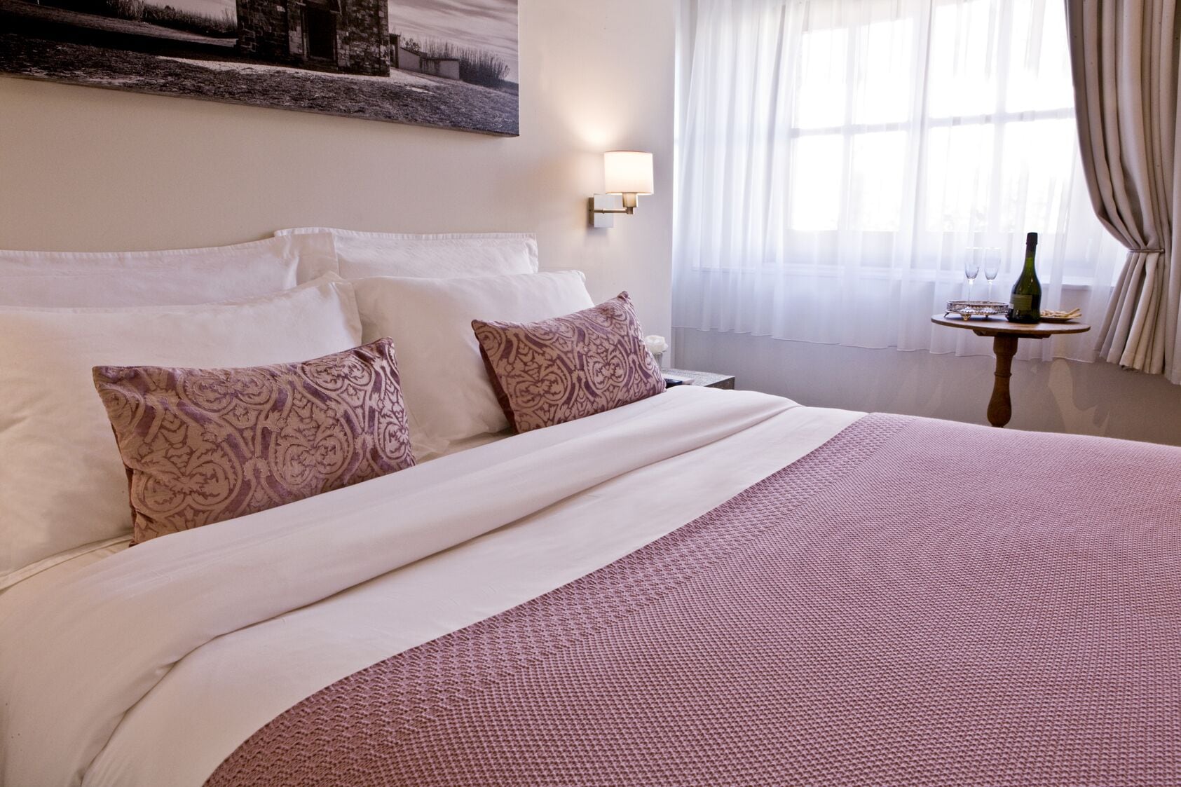 A neatly made bed with patterned pillows, a bedside table with a bottle and glasses, and a window with sheer curtains.