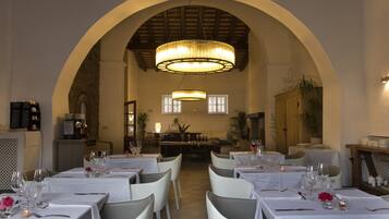 A restaurant with white tablecloths, chairs, and a large chandelier. Tables are set with wine glasses and cutlery.