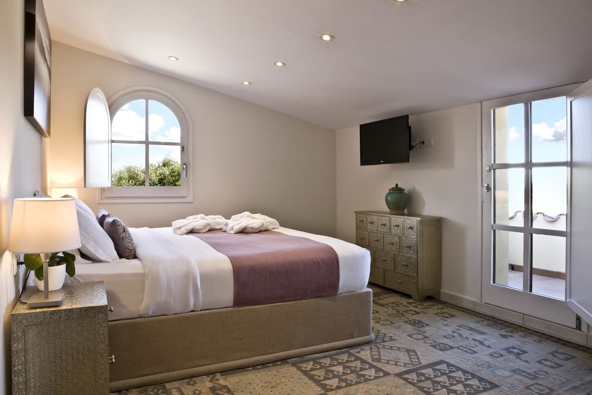 Ein Schlafzimmer mit Bett, Nachttisch, Kommode, Fenster und einem an der Wand befestigten Fernseher.