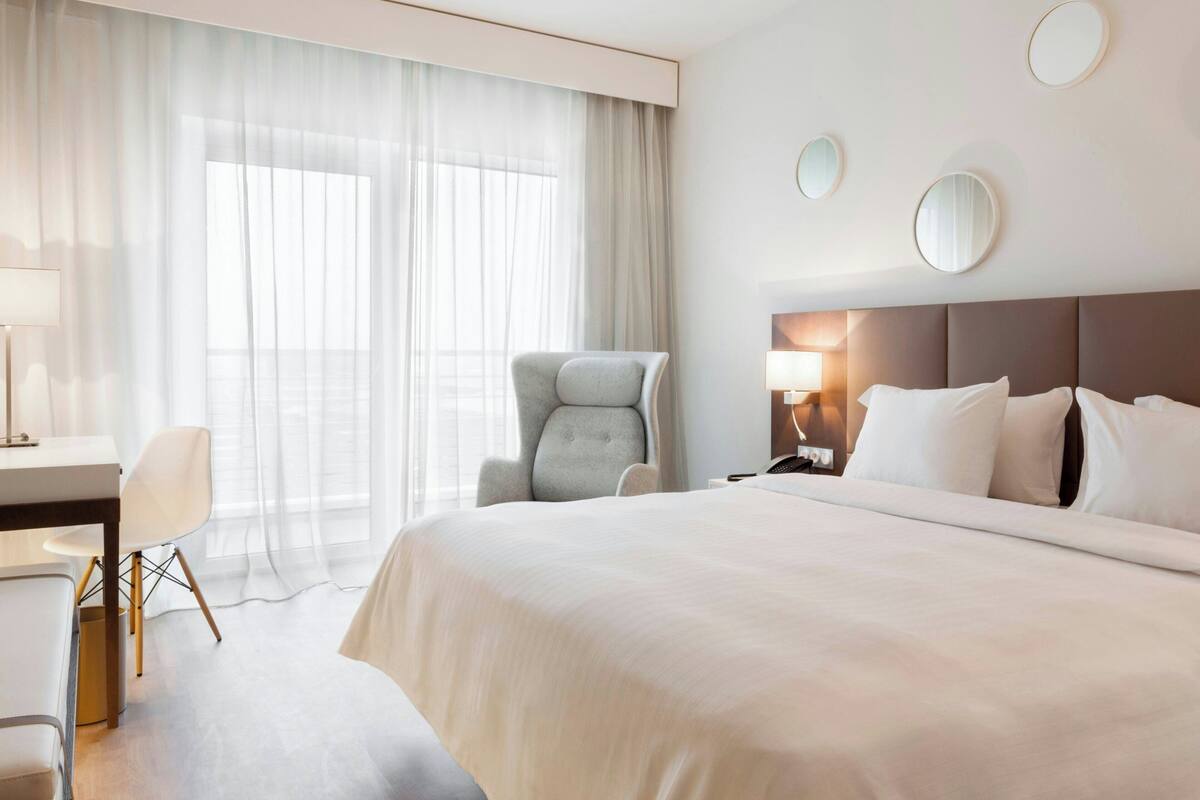 Une chambre d’hôtel avec un grand lit, un bureau avec une chaise, une lampe et une fenêtre avec des rideaux légers.