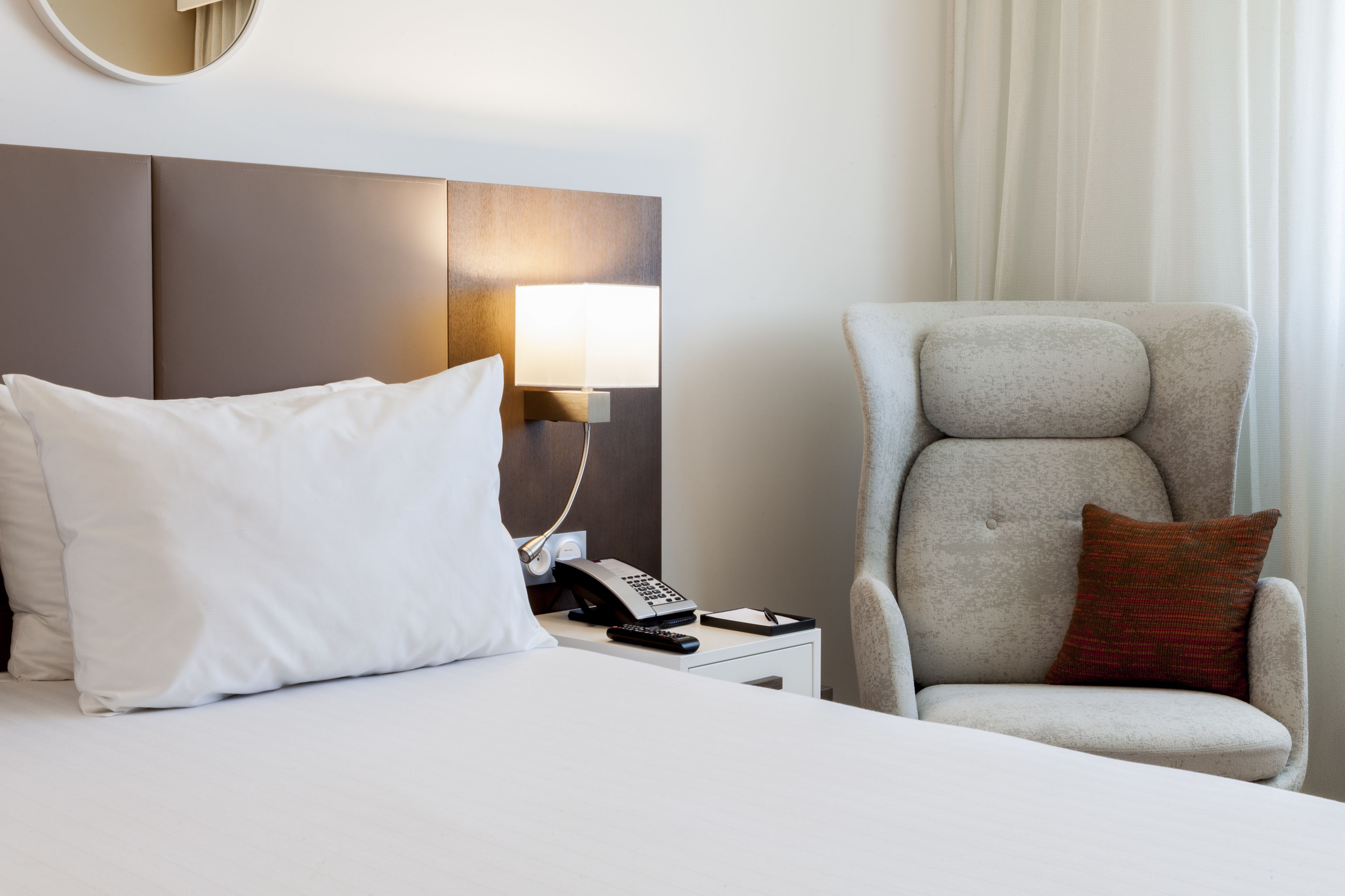 Une chambre d’hôtel avec un lit, une table de chevet avec une lampe, un téléphone et une chaise avec un coussin.