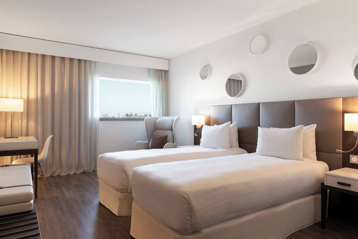 Une chambre d’hôtel avec deux lits, un bureau, une chaise, une fenêtre avec des rideaux et une vue sur la ville.