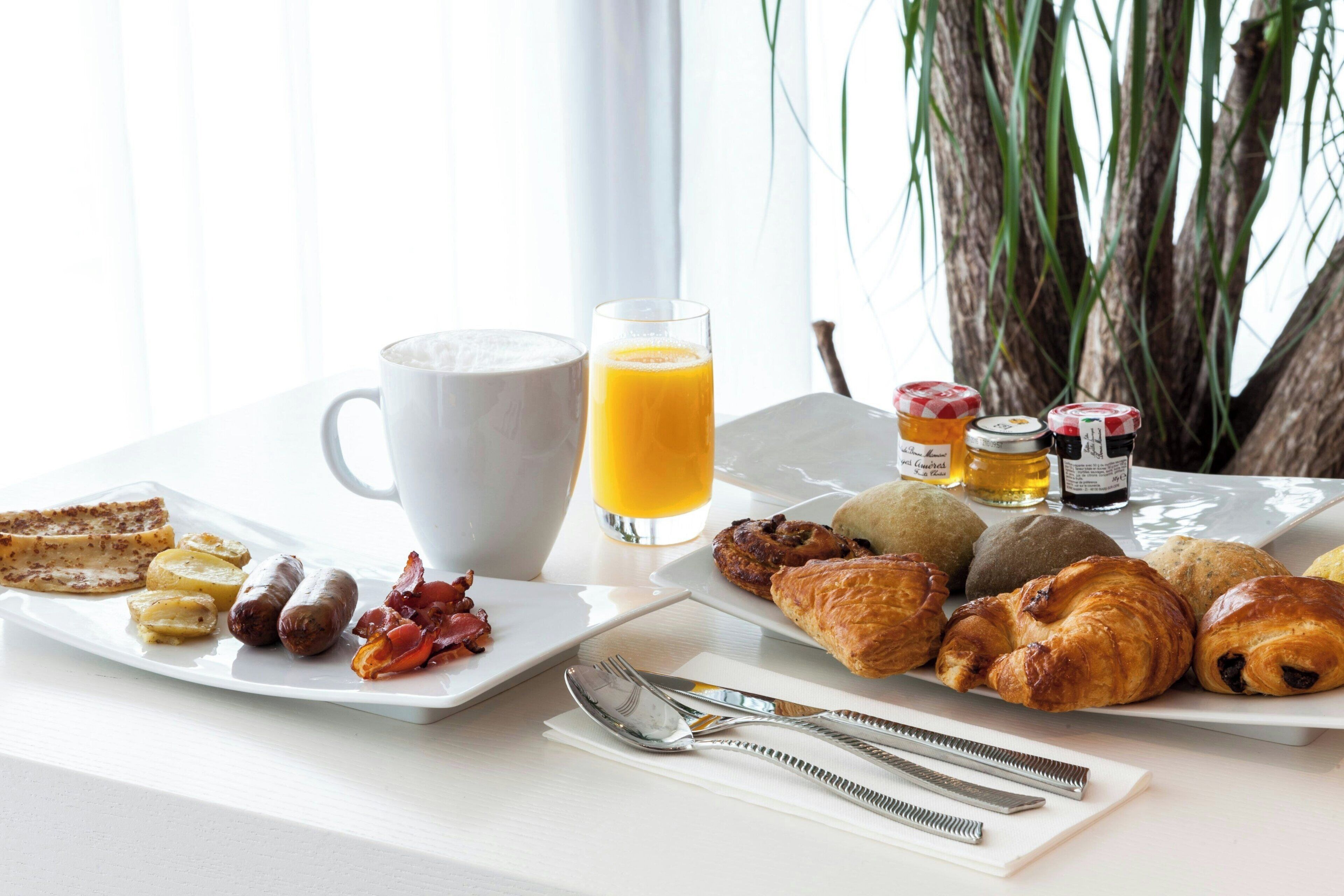 Een ontbijt met toast, worstjes, spek, eieren en gebak op een wit bord, een glas sinaasappelsap en een kop koffie.
