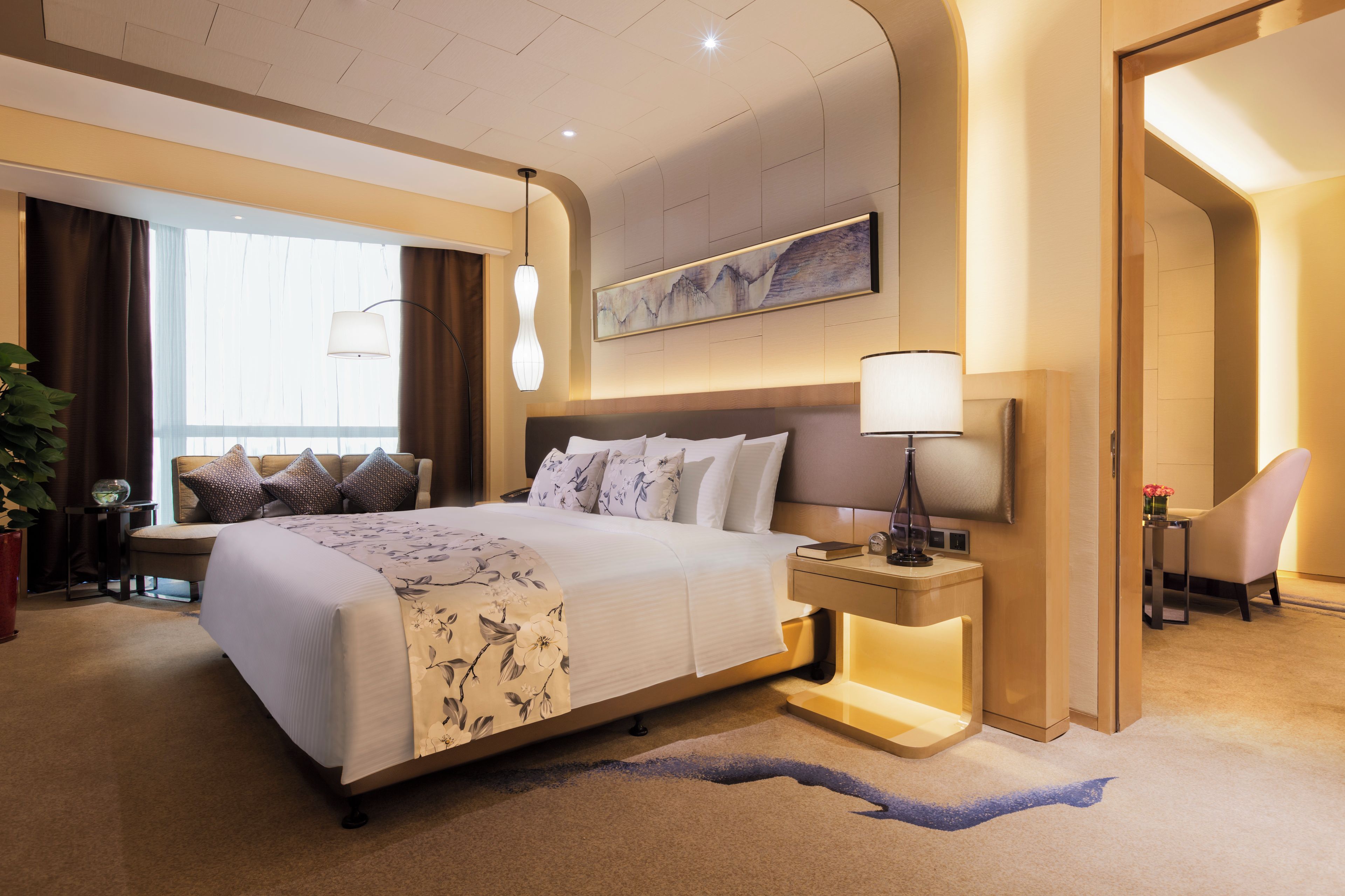 Ein Hotelzimmer mit einem großen Bett, Nachttischen, einem Schreibtisch, einem Sessel, einer Topfpflanze und einem Gemälde an der Wand.