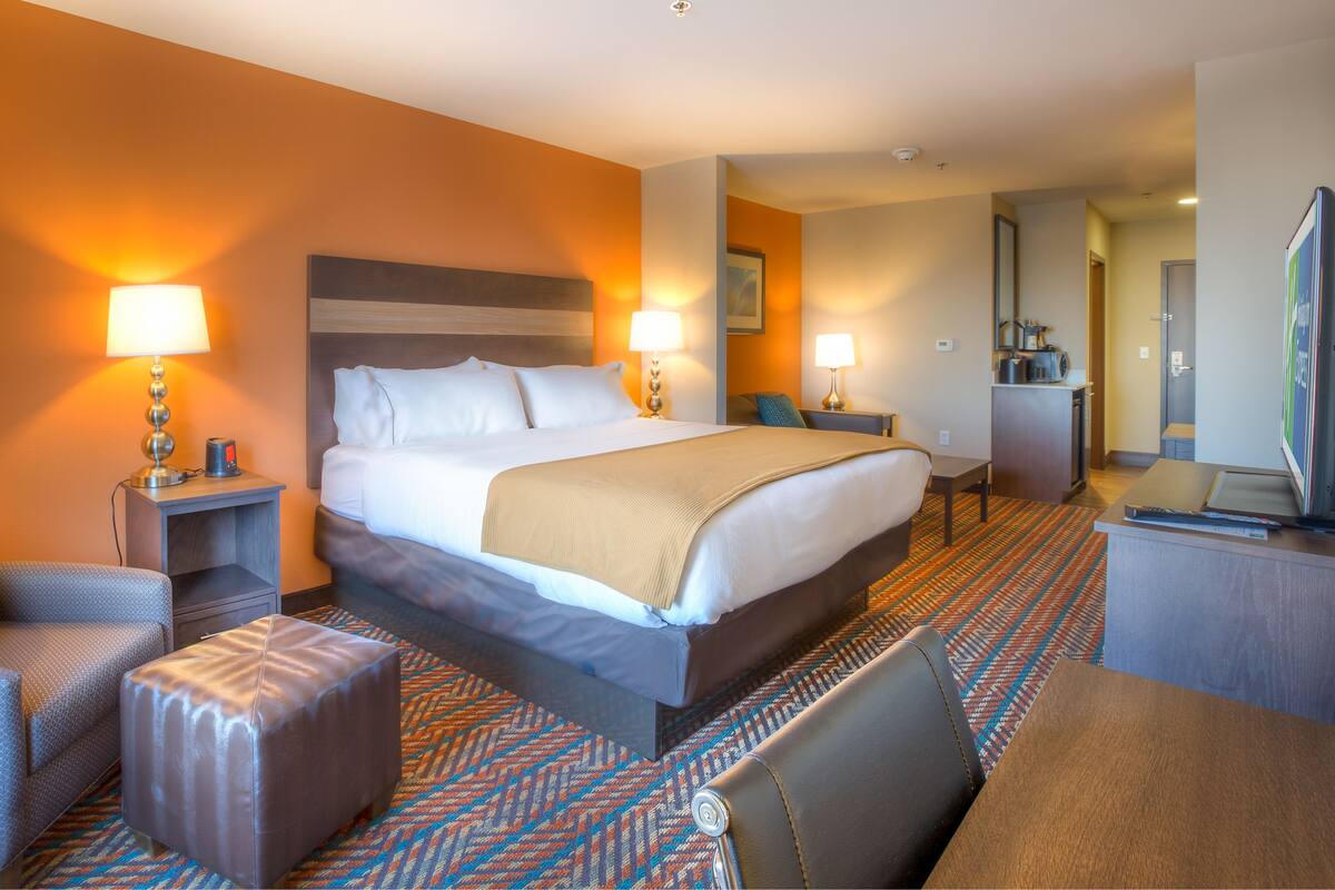 A hotel room with a large bed, two bedside tables with lamps, a sofa, a small ottoman, a desk, and a television.
