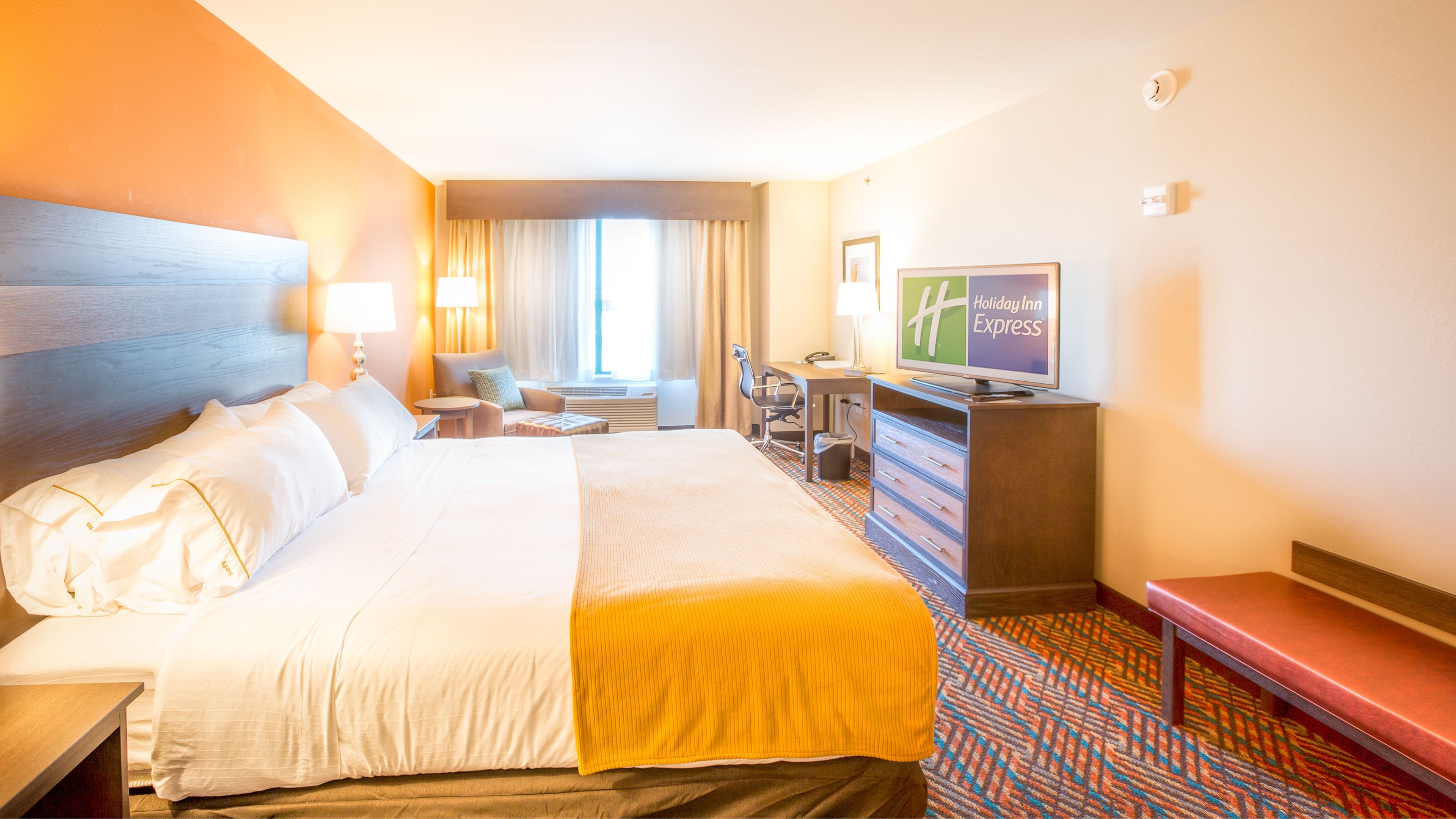 A hotel room with a large bed, a desk with a computer, a television, and a seating area with a chair and a small table.