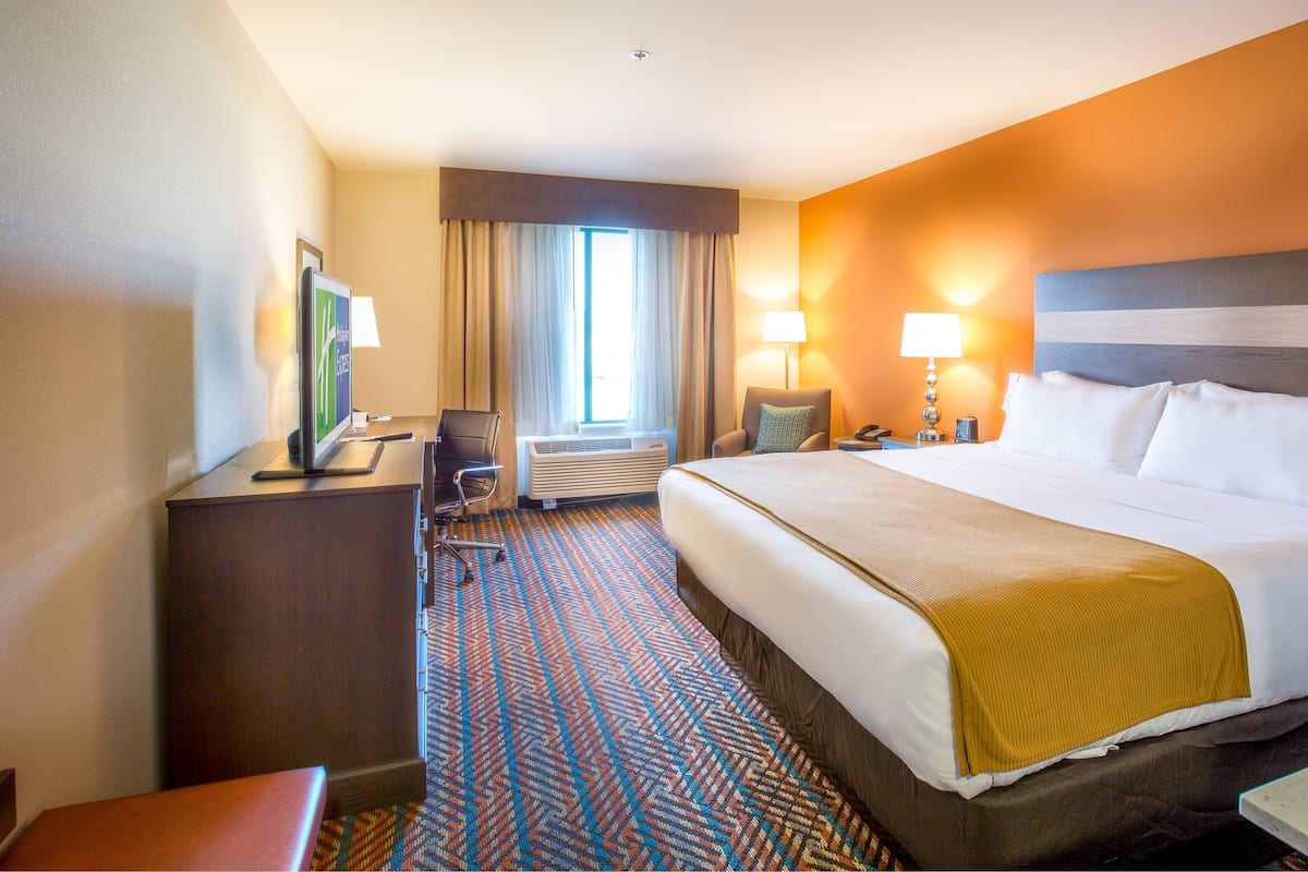 A hotel room with a large bed, a desk with a computer, a chair, and a window with curtains.