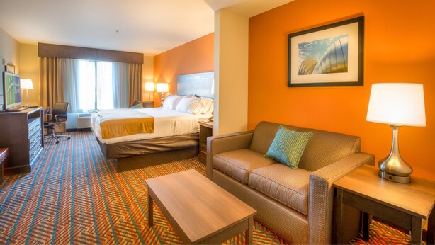 A hotel room with a large bed, a sofa, a desk, and a chair. There is a painting on the wall and a window with curtains. - Holiday Inn Express Wichita South by IHG (Wichita)