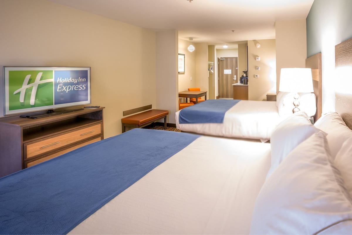A hotel room with two beds, a television displaying 'Holiday Inn Express', a desk with a chair, and a bathroom visible in the background.
