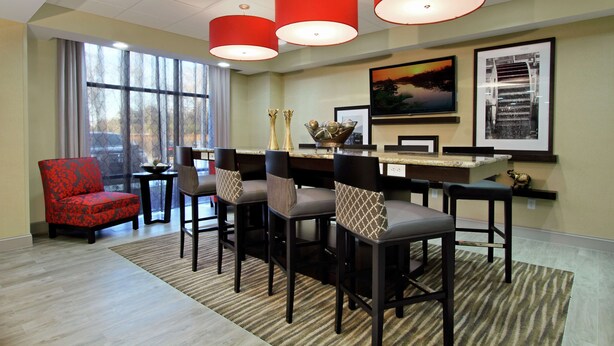 A hotel room with a dining area featuring a bar and multiple bar stools. There is a red upholstered chair and a small table with decorative items. - Hampton Inn Richland (Richland)