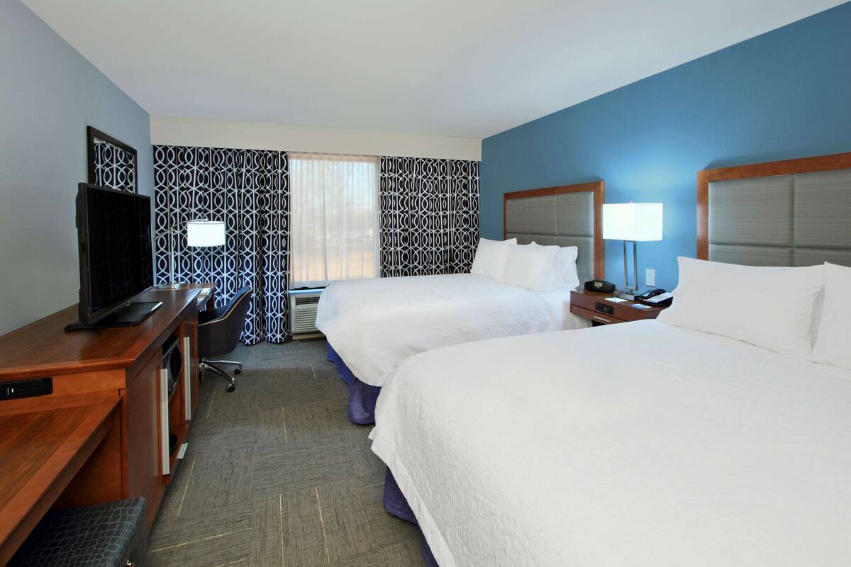 A hotel room with two beds, a desk with a television, a lamp, and a window with patterned curtains.