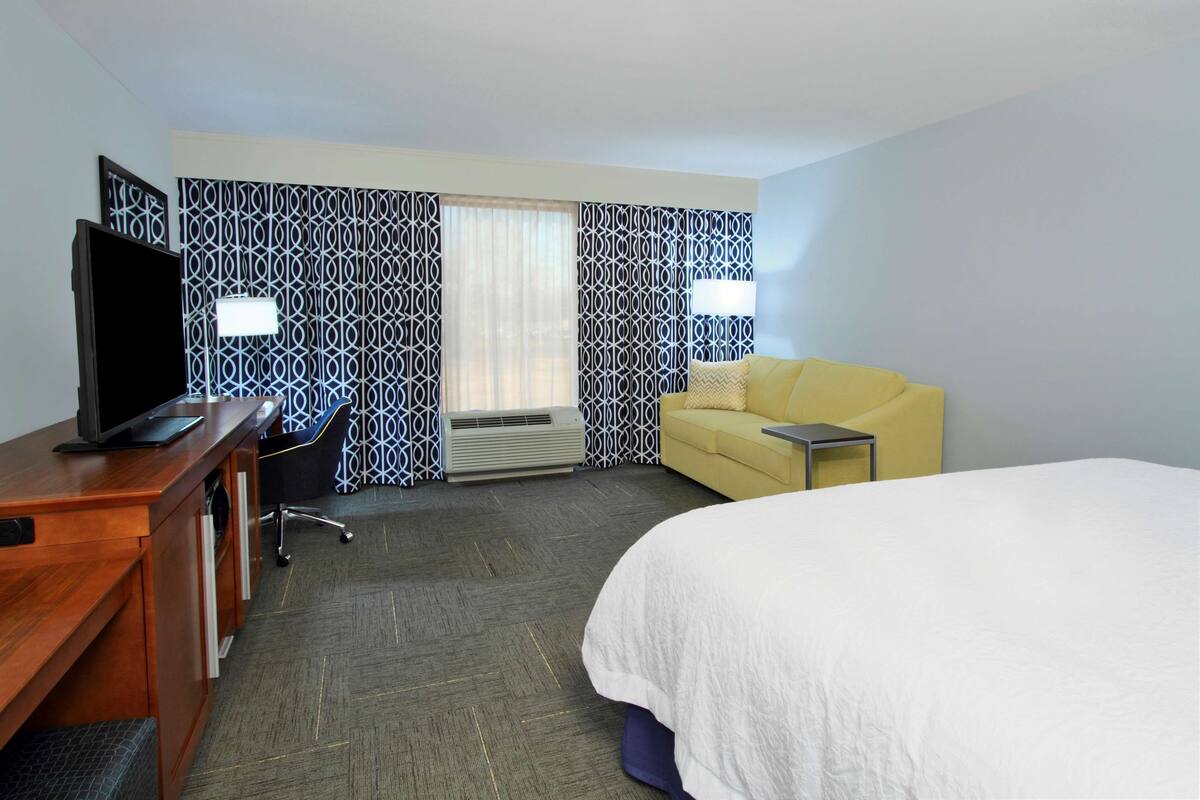 A hotel room with a bed, a desk with a TV, a yellow sofa, and patterned curtains.