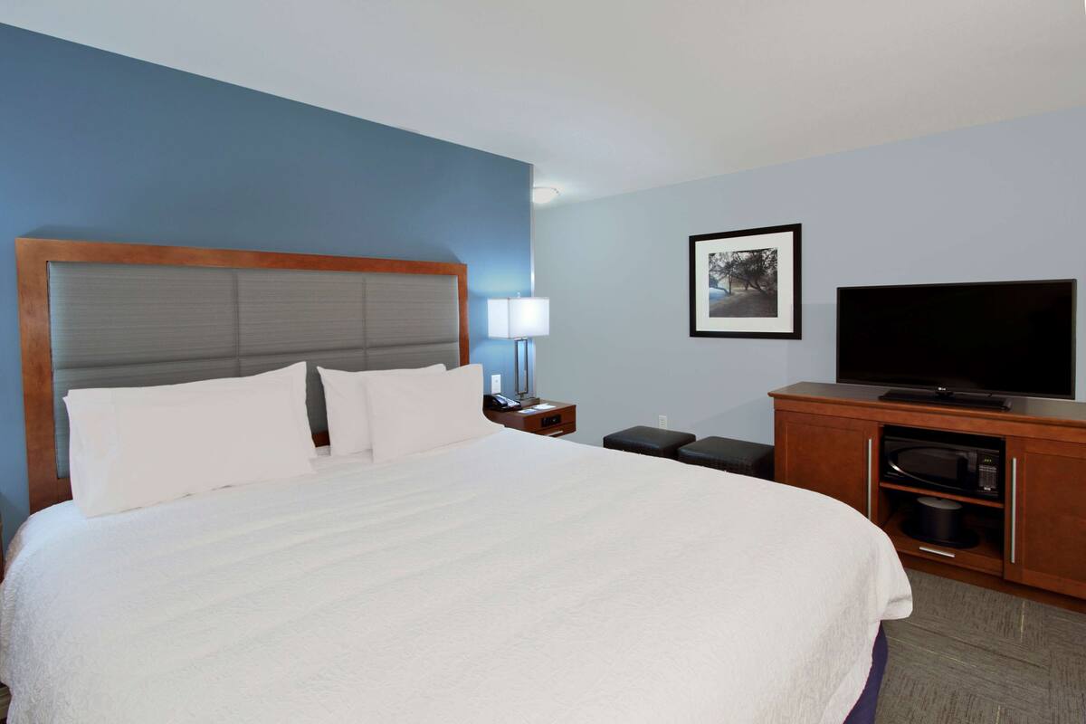 A hotel room with a large bed, a television, a nightstand with a lamp, and a microwave in a wooden cabinet.