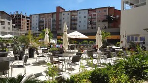 Area patio all'aperto con tavoli e sedie, circondata da piante in vaso e ombrelloni.