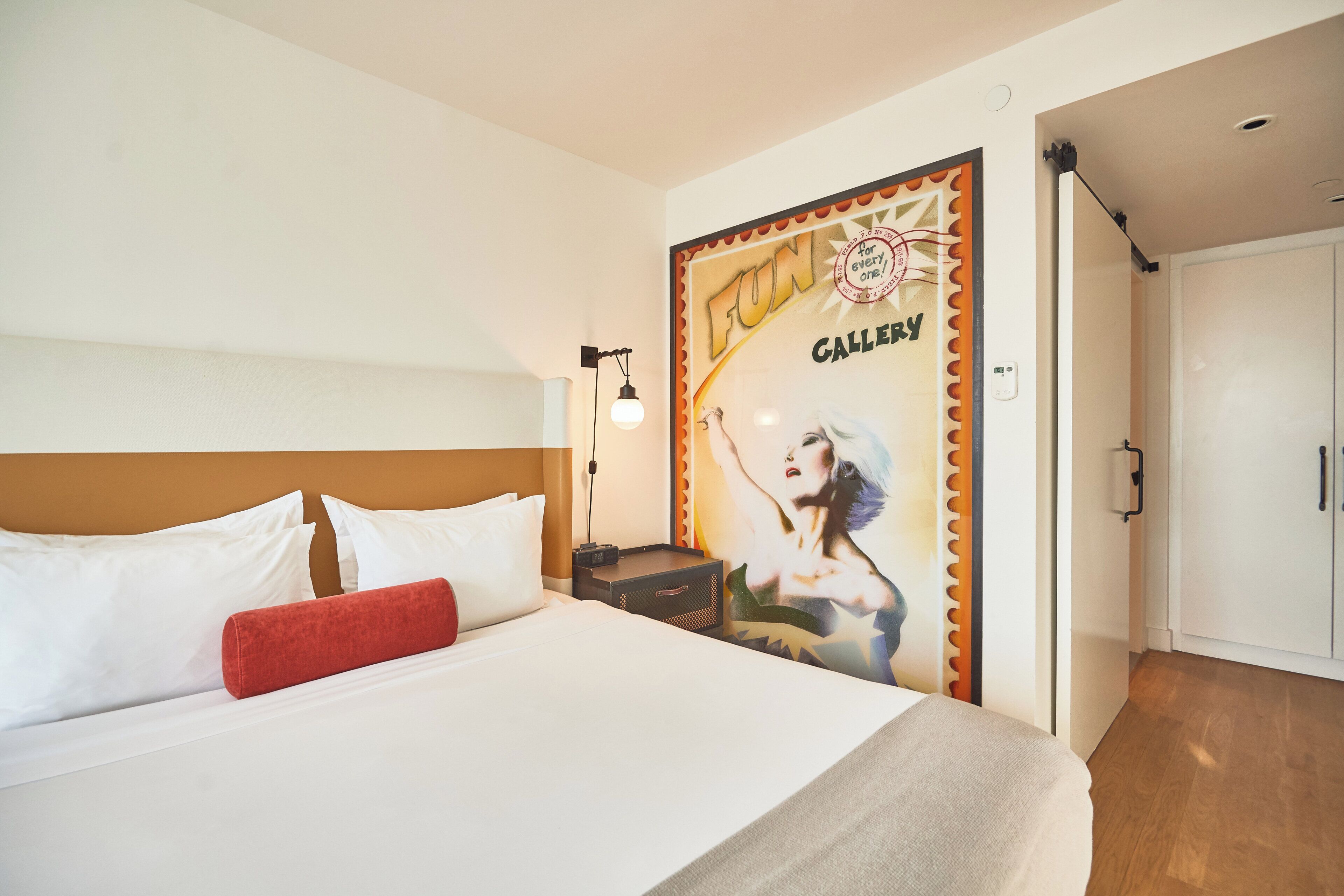 A hotel room with a bed, a nightstand, a wall-mounted lamp, and a poster on the wall.