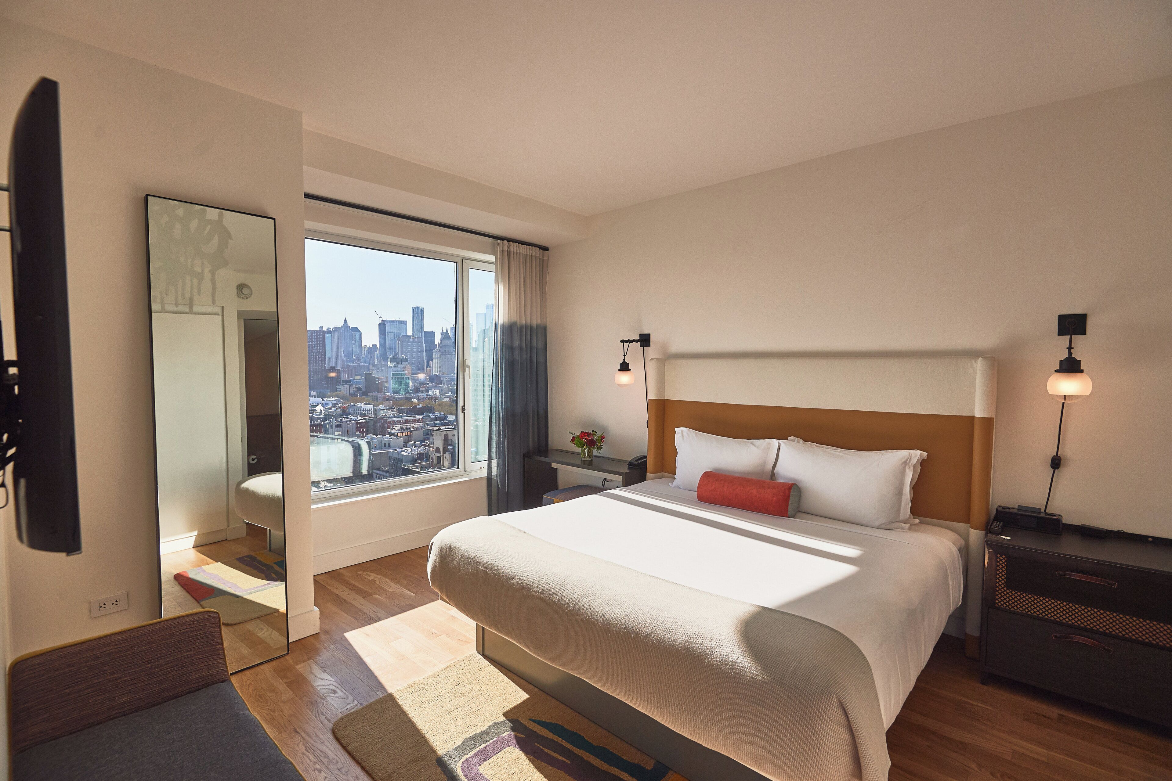 A hotel room with a large bed, a nightstand, a lamp, and a view of the city through a window.