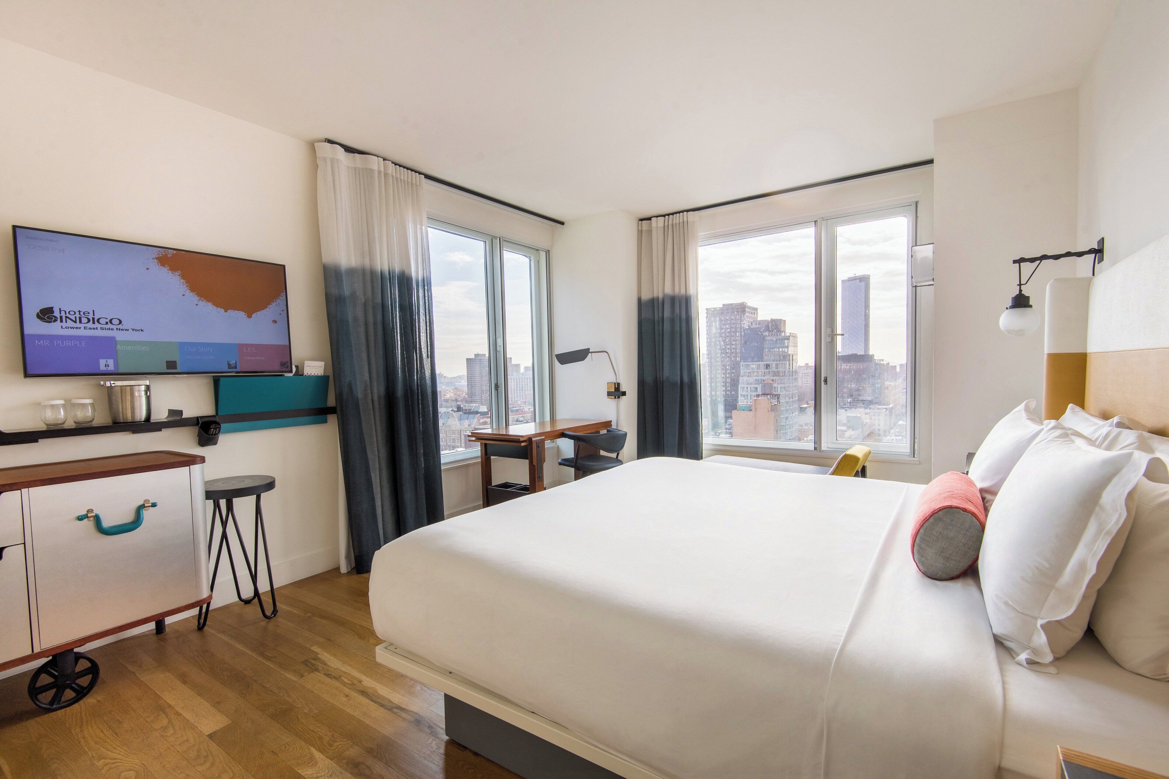 A hotel room with a bed, a desk, a chair, a television, and a view of the cityscape.