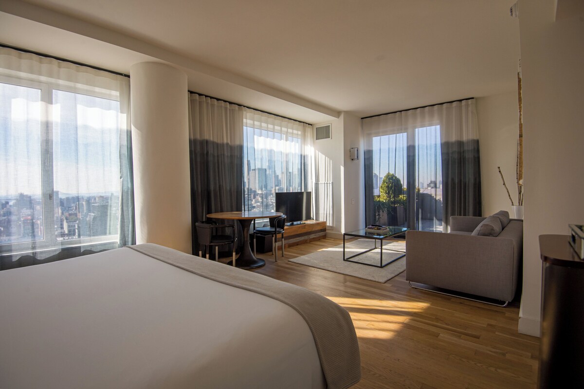 Una habitación de hotel moderna con una cama grande, un sofá, una mesa de comedor y un televisor. La habitación tiene ventanas amplias con cortinas, que ofrecen vistas a la ciudad.