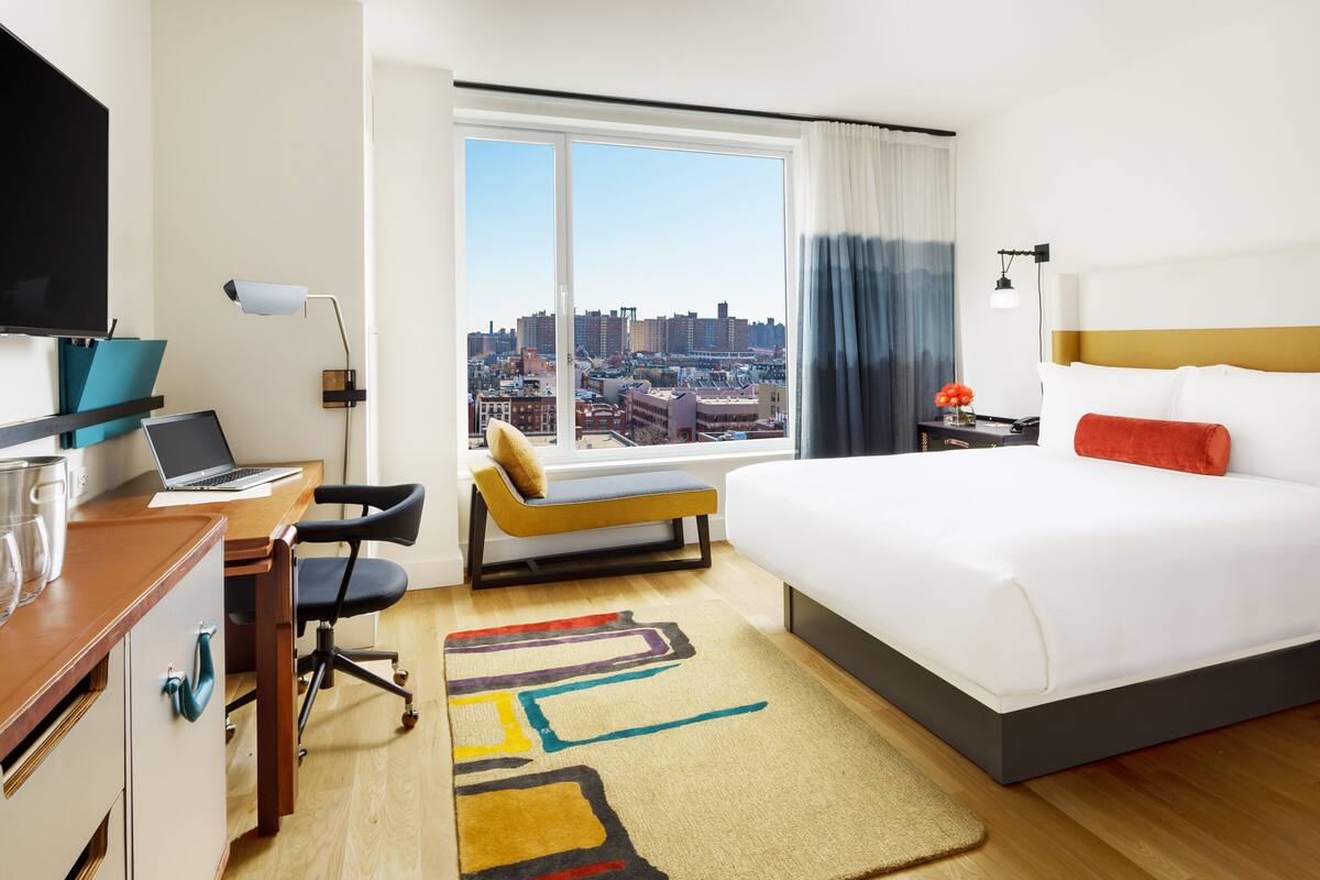 A hotel room with a large bed, a desk with a laptop, a chair, a TV, and a view of the city.