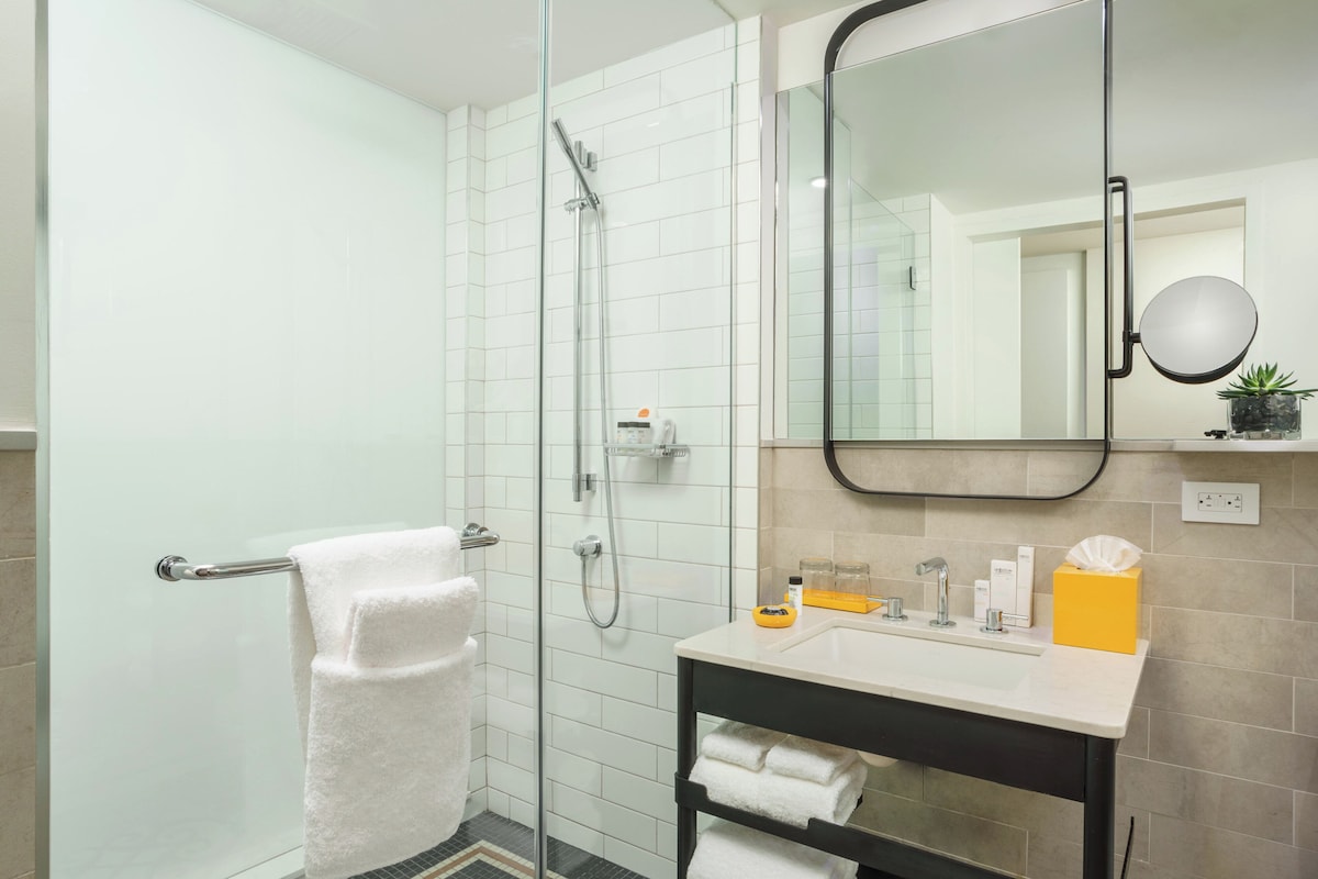 Un baño moderno con ducha de vidrio, toallero blanco, lavamanos con artículos de aseo y un espejo.