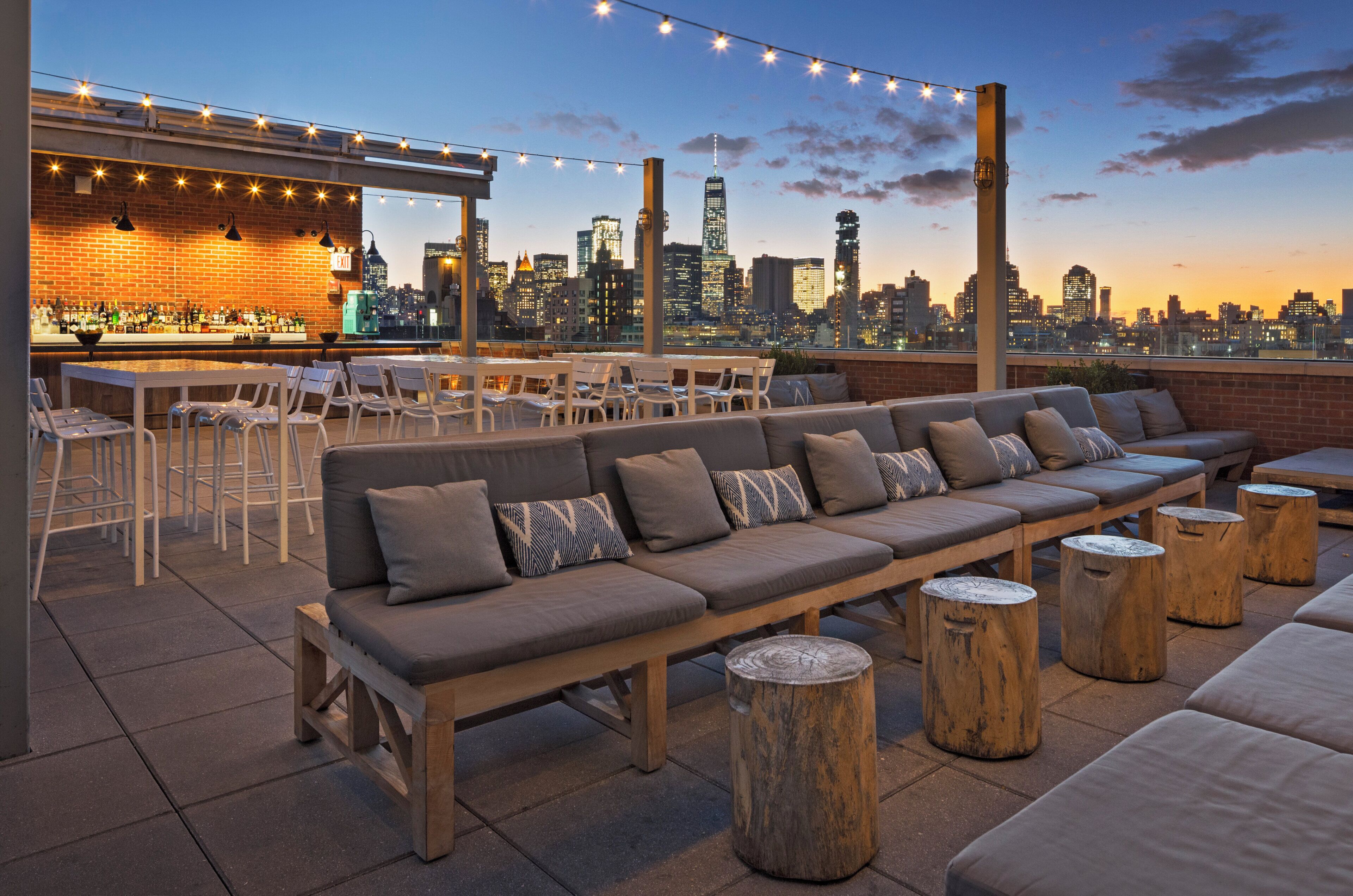 An outdoor seating area with a city skyline view, a bar with a brick wall, and string lights.