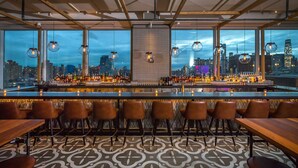 Een bar met uitzicht over de stad, met een lange toonbank, meerdere hangende lampen en een vloer met patroon.