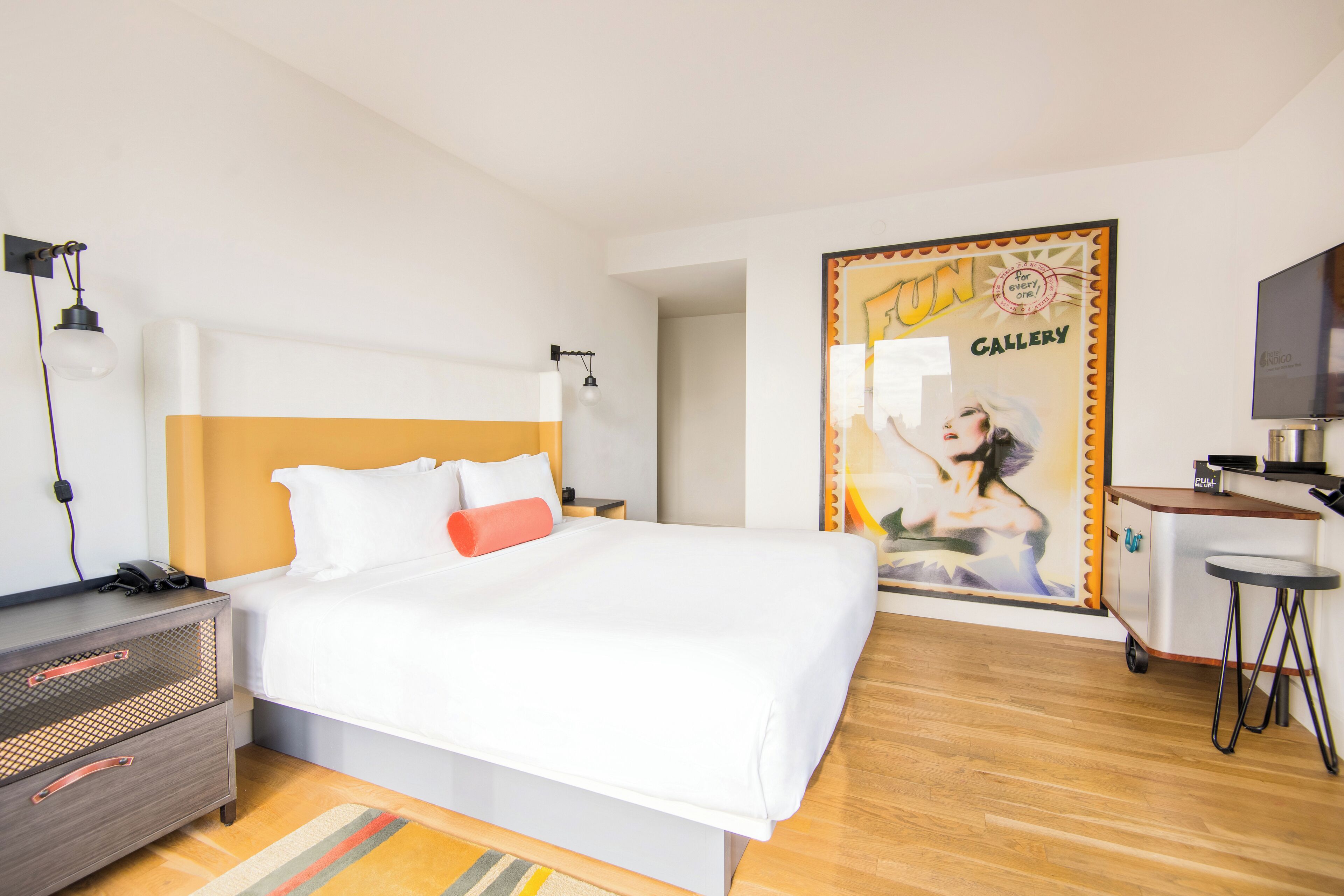 A hotel room with a bed, a desk, a TV, and a poster on the wall.