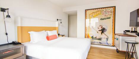 A hotel room with a bed, a desk, a TV, and a poster on the wall.