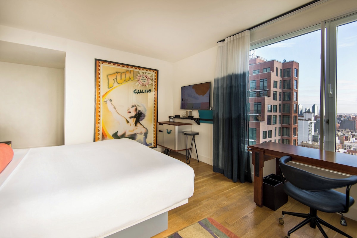 Habitación de hotel con cama, escritorio, silla y un amplio ventanal con vista a la ciudad.